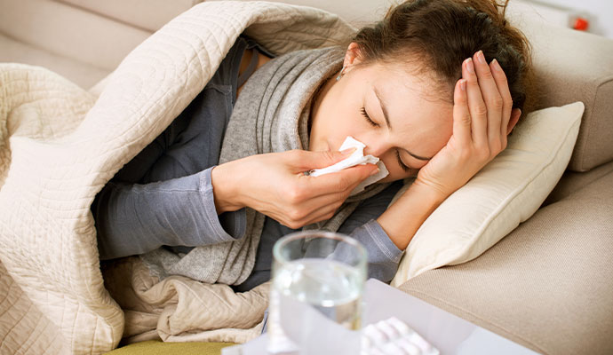 A young sick woman lying on a beige sofa wrapped in a blanket, holding a tissue to her nose