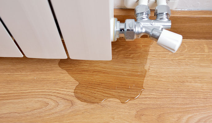 A puddle of water on the floor underneath a white radiator's chrome control valve